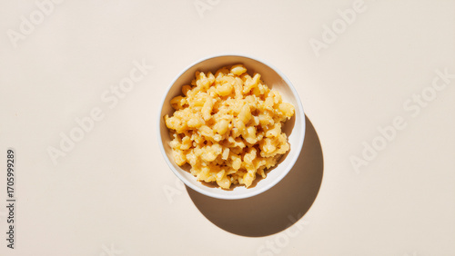 Mac and cheese in a plain white bowl on a light ivory background, clean top-down studio shot with glossy cheese highlights and a soft halo shadow