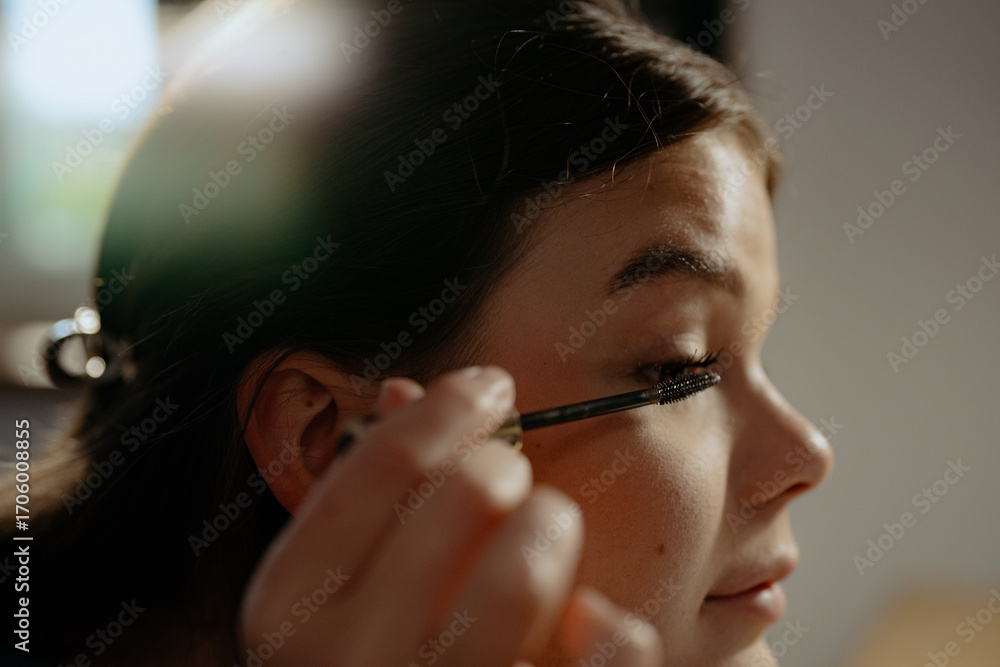 Obraz premium A young woman carefully applies mascara to her eyelashes while sitting in a bright room. Soft sunlight filters through a window, enhancing her focused expression and room ambiance.