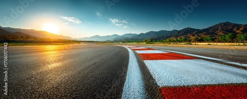 Asphalt racing track leads to distant mountains, lit by warm sunlight under a blue sky with sparse clouds. Perspective is low angle
