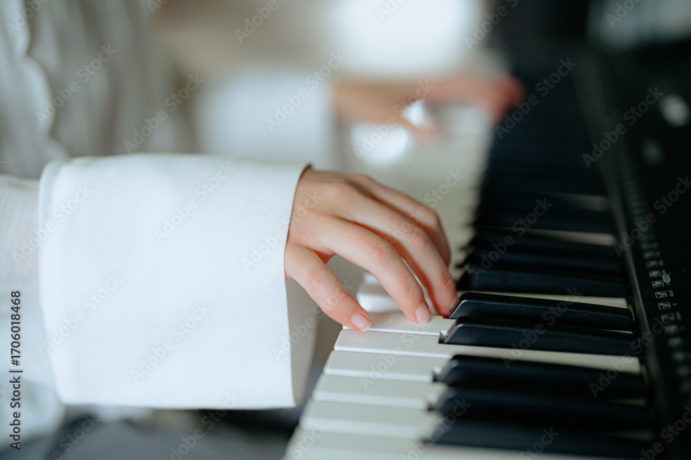 Obraz premium A person in a white shirt gracefully plays the piano, their fingers moving over the black and white keys. The bright room adds a warm atmosphere to the musical activity.