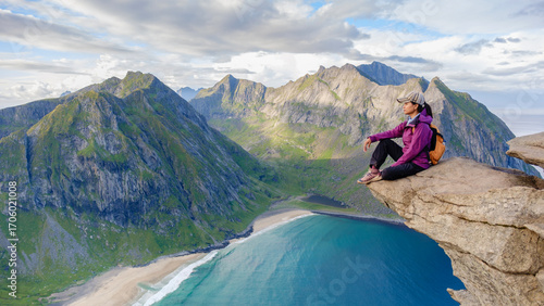 Photos A hiker rests at the edge of a rocky cliff on Ryten, admiring the stunning panorama of Kvalvika Beach and the rugged Lofoten landscape during a clear day