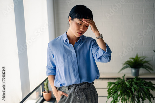 Tired Asian woman standing indoors with hand on forehead, expressing stress, fatigue, or headache, representing exhaustion and emotional struggle