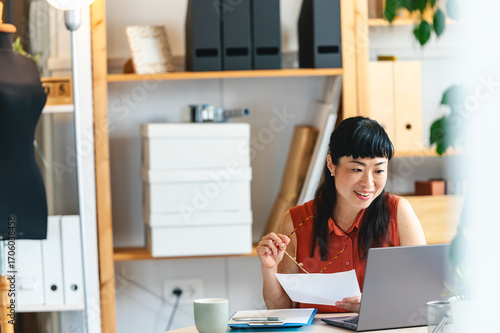 Female small business entrepreneur working at desk with laptop, holding document and looking thoughtful while managing tasks in modern office workspace