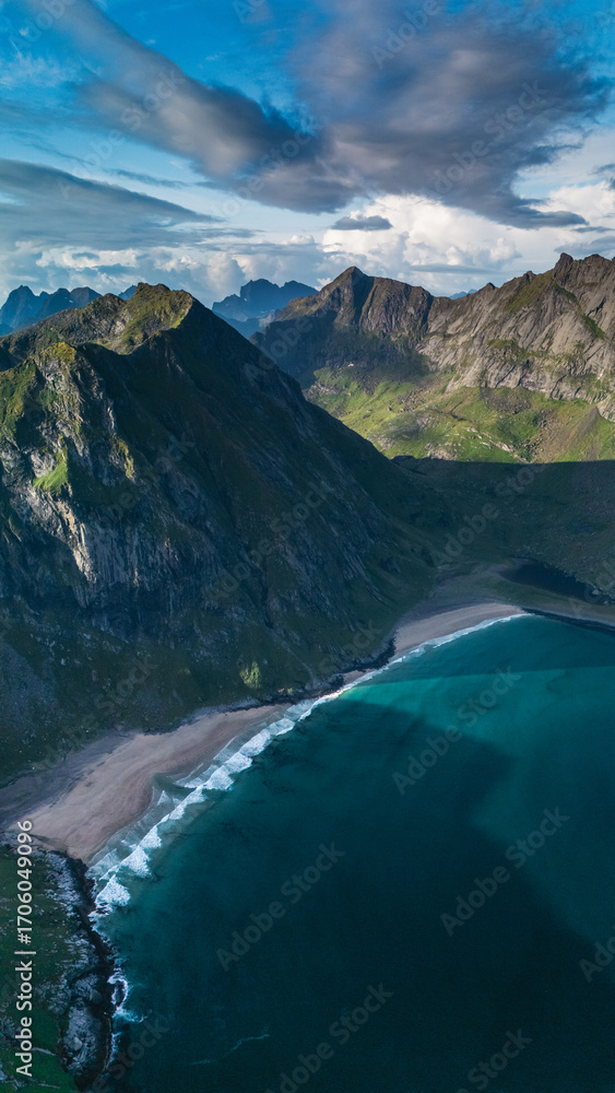 Fototapeta premium Stunning aerial perspective of Ryten Kvalvika beach trail, showcasing lush green mountains, vibrant blue ocean waves, and sunny skies. Ideal for nature lovers and outdoor adventure enthusiasts.