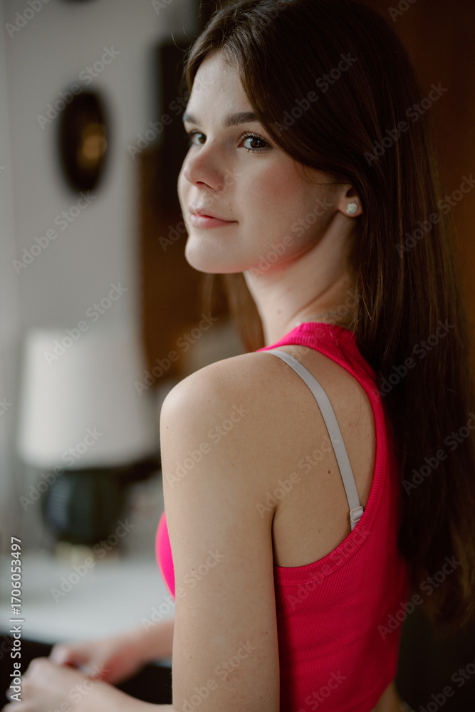 Fototapeta premium A young woman with long brown hair is standing casually in a bright indoor space. She wears a pink top and looks over her shoulder with a confident expression, enjoying a moment of relaxation.