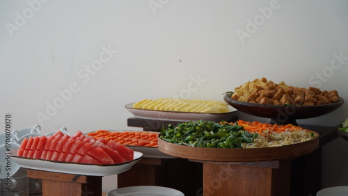 A colorful buffet spread featuring large platters of sliced watermelon, carrots, and various chopped vegetables next to fried food, presented on wooden risers against a plain wall.