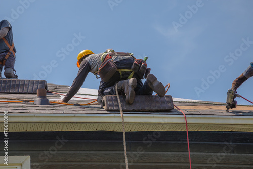 Construction Workers with Safety Equipment Installing Asphalt Shingles on Residential Roof