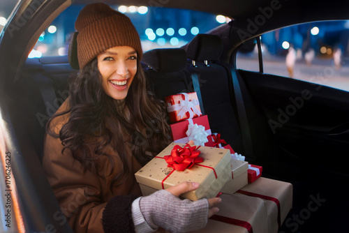 Excited woman preparing for winter holidays, sitting with Xmas gift boxes on back seat of car and smiling at camera