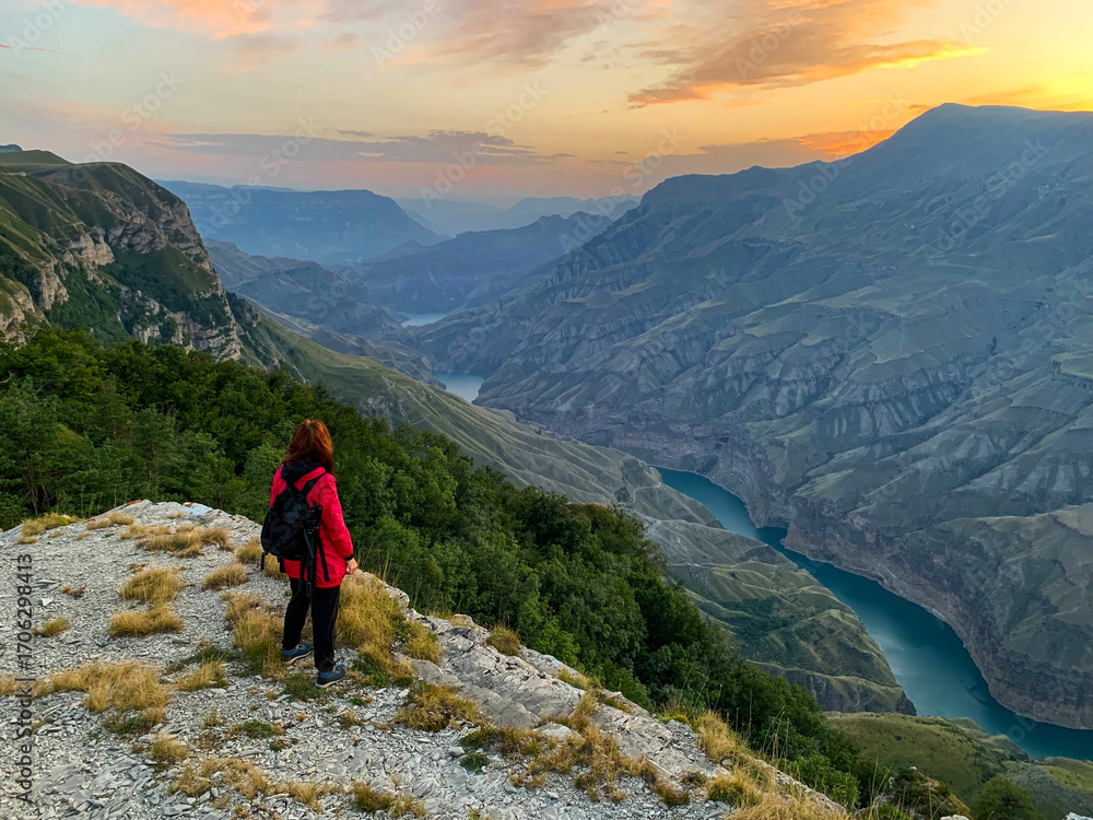 Fototapeta premium A woman in a red jacket stands on a rocky cliff above a lush valley and a winding river at sunset