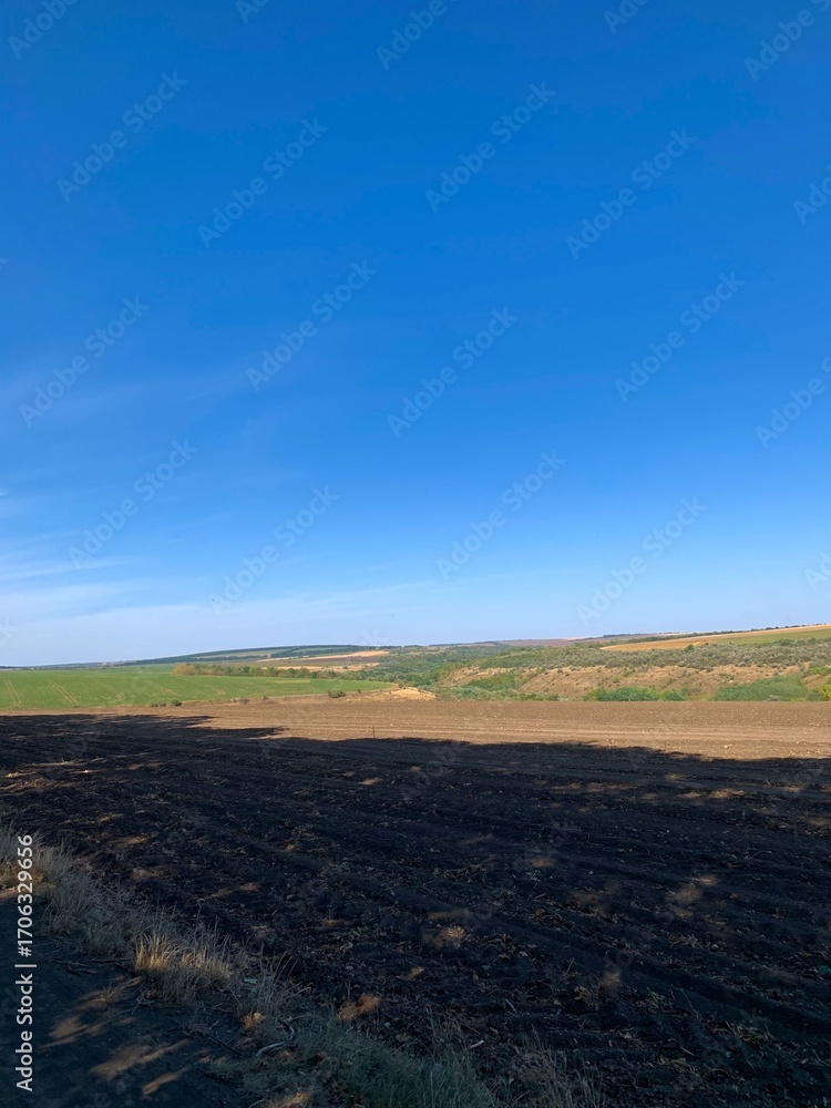 Fototapeta premium Wide rural landscape with burnt soil in the foreground, green fields and hills under a clear blue sky. Concept of agriculture, nature, and ecological contrast.