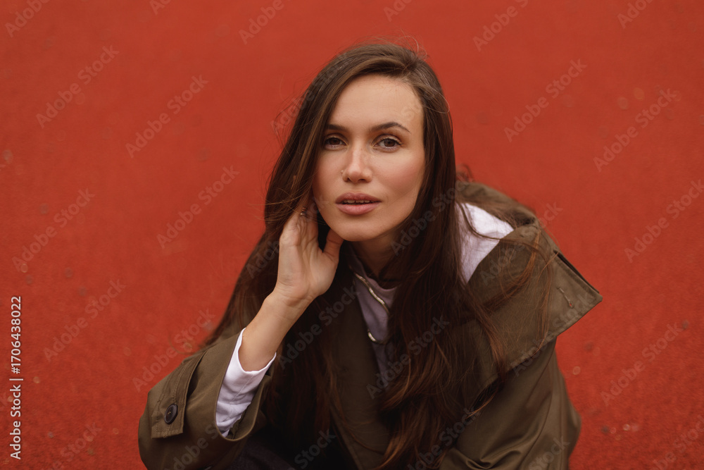 Naklejka premium Portrait of elegant woman sitting outdoor, hold her head, look sensual and dreaming, look at camera. Girl wear raincoat.
