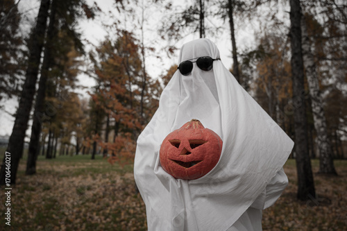 Woman in white sheet holding jack o'lantern against autumn forest background. Halloween costume. 