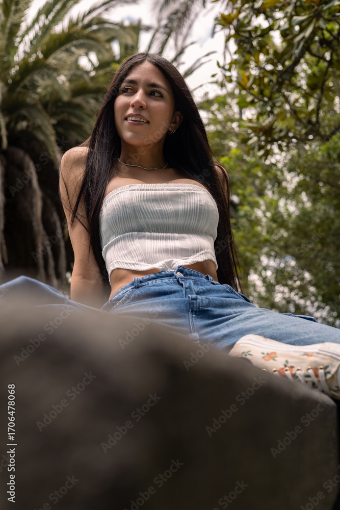 Obraz premium Beautiful young woman with long dark hair sits gracefully on a rustic stone bench in a sunlit garden. Wearing denim jeans and a white crop top, she exudes charm, elegance, and relaxed confidence while