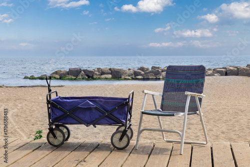A four wheel beach wagon by a folding beach chair  on a board walk by the water.