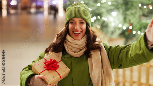 Happy young European woman with gift boxes in hands posing outside on Christmas Eve, taking selfie with decorated festive background. Concept of shopping and holidays