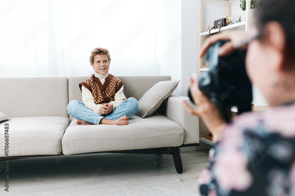 Obraz premium Authentic play scene featuring a young boy sitting cross legged on a sofa, dressed in playful school style, as a photographer captures candid moments at home.