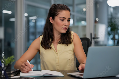 Focused Professional Woman Taking Notes While Working on Laptop in Modern Office