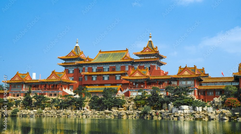 Naklejka premium Traditional Chinese temple in Penglai, Shandong, with red pillars, golden roofs, and ornate details, surrounded by lush greenery and a serene natural setting.