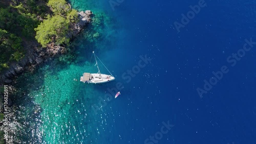 A stunning top down drone video of a sailboat anchored in a secluded cove with crystal clear turquoise water next to a rocky and forested coastline a paradise for sailors
