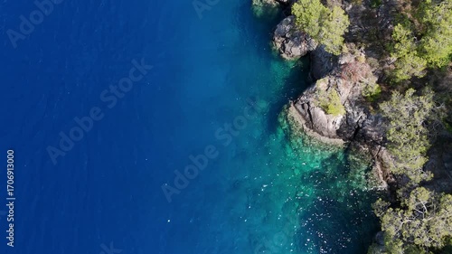 A top down aerial video of a deep blue sea next to a rocky coastline with clear turquoise water revealing the sea floor a stunning and contrasting natural border