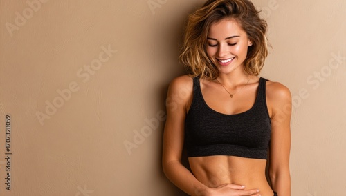 Smiling fit woman in athletic wear leans against a neutral wall, showcasing a toned physique