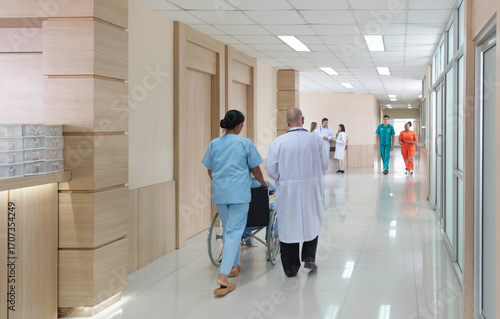 Two health care professional walk with a patient in a wheelchair down a well-lit hospital corridor, where multiple staff member are attending to their duty.
