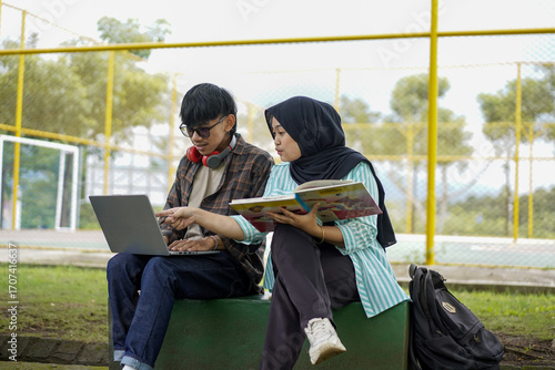 Two students studying outdoors, using a laptop and a book, likely collaborating or working on a project together, at a park or campus setting
