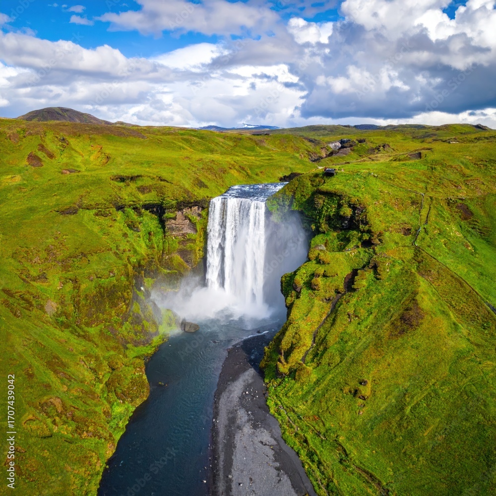 Obraz premium Aerial view of a waterfall crashing down into a rocky riverbed amidst vibrant green hills, under a partially cloudy sky
