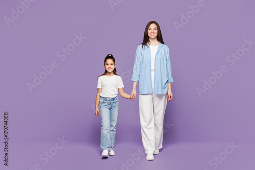 Full body young woman wear blue clothes have fun with child baby girl 8-9 years old. Mommy little kid daughter hold hands walk go look camera isolated on plain purple background. Mother's Day concept.