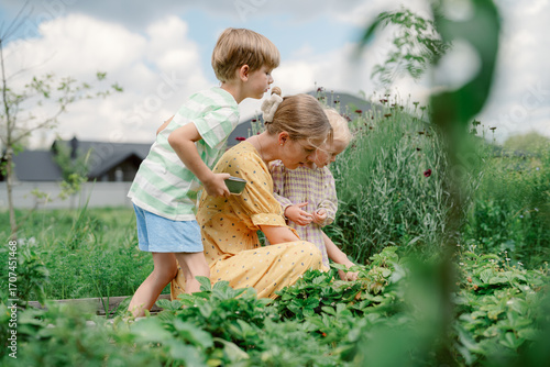 A woman kneels among vibrant plants while two children help her pick fresh strawberries. The garden is lush and green under a bright sky, showcasing blossoming flowers and vegetables.