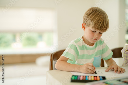 Фототапет Young boy focused on drawing with colorful markers on a white sheet at home