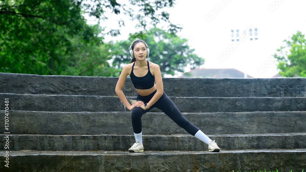 Fototapeta premium Female athlete wearing headphones, warming up before workout in urban park