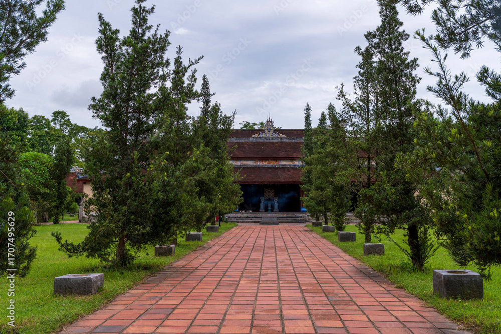 Obraz premium A brick pathway leads symmetrically toward a traditional temple building, flanked by manicured cypress trees and green lawns, creating a peaceful and spiritual atmosphere.