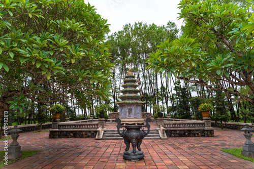 A multi-tiered stupa and a large bronze urn stand on a red brick courtyard, framed by beautiful frangipani trees, creating a serene and spiritual scene.