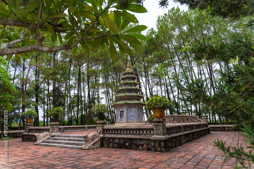 A multi-tiered stupa and a large bronze urn stand on a red brick courtyard, framed by beautiful frangipani trees, creating a serene and spiritual scene.