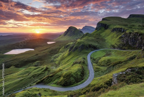Winding road curves through lush, green hills under a vibrant sunset. Peaks rise majestically, with a dramatic sky overhead
