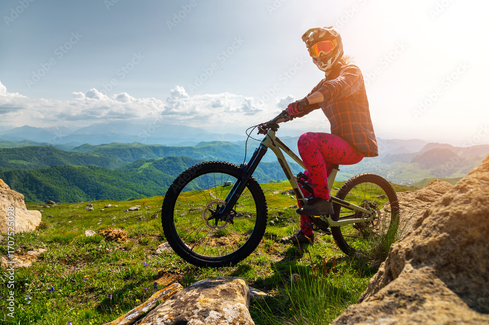 Naklejka premium Tourist with bicycle enjoying valley view from mountain top. Male cyclist riding mountain bike outdoors. Male tourist standing on grassy path in mountains wearing helmet