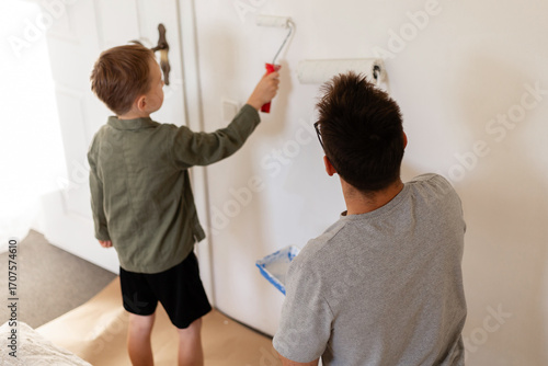 A man and his young son are painting the walls of their bedroom. They are whitewashing the walls as part of a home improvement project. The boy actively helps his father