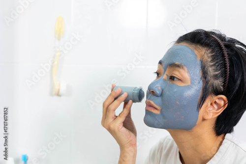 Asian woman applies clay facial mask in front of bathroom mirror. Skincare routine promotes clean skin and natural beauty. A refreshing beauty treatment for detoxifying and rejuvenating the face.
