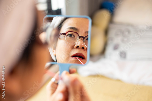 Beautiful Asian woman applies lipstick in front of handheld mirror. Close-up reflection shows candid beauty routine of self care, natural skin, and confident expression during personal makeup moment.