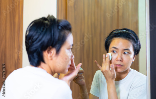 Young Asian woman gently cleanses her face with a cotton pad and cleanser in bedroom. Skincare routine focuses on makeup removal, self care, and achieving a clean, healthy face with mirror reflection.