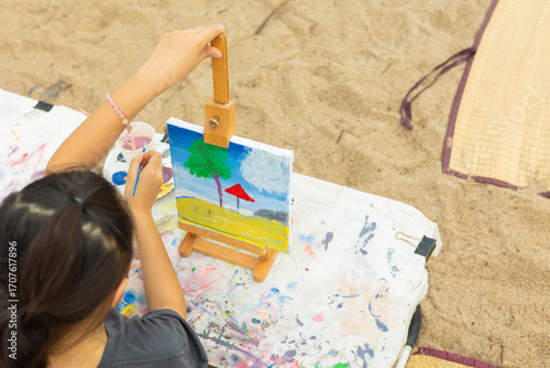 Close-up side view of a young girl focused on her painting at an easel outdoors. This shows the intense concentration and creativity of a child engaged in an artistic hobby or art class.