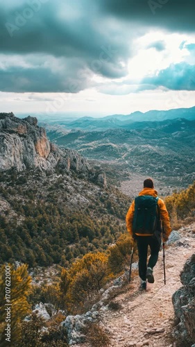 A man wearing a yellow jacket and a blue backpack is walking on a trail. He is carrying two walking sticks
