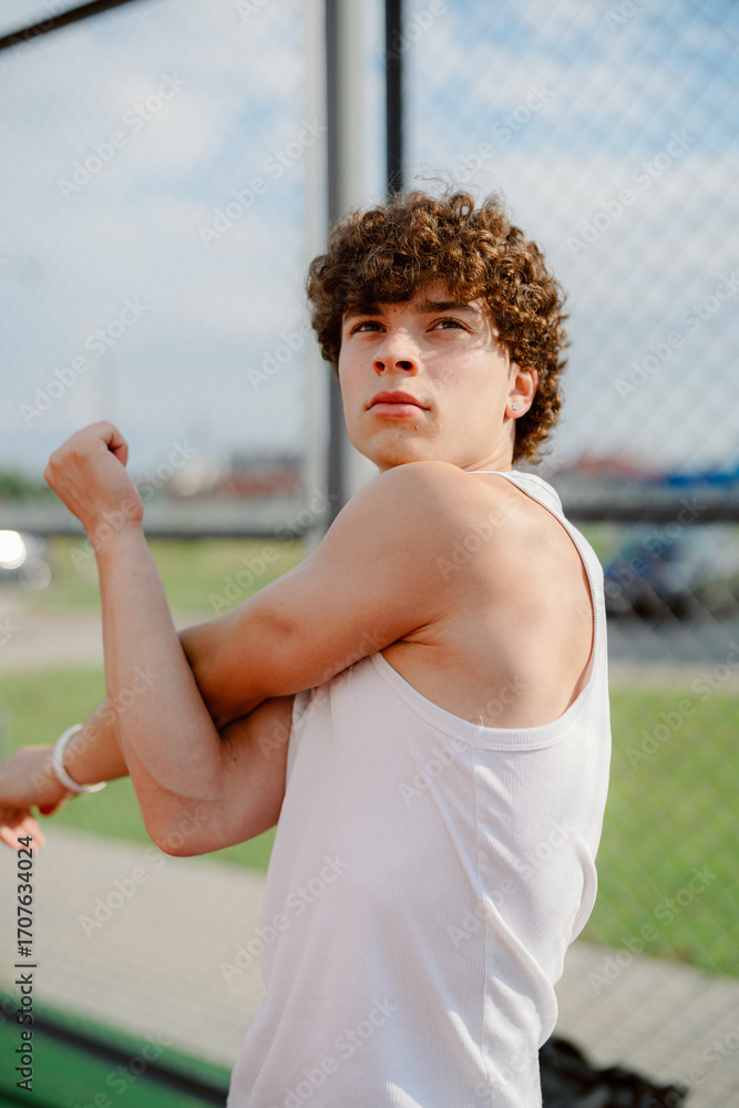 Obraz premium A young male athlete prepares for his practice by stretching his arm in a sports facility. The setting features a green field and a clear blue sky, indicating daytime.