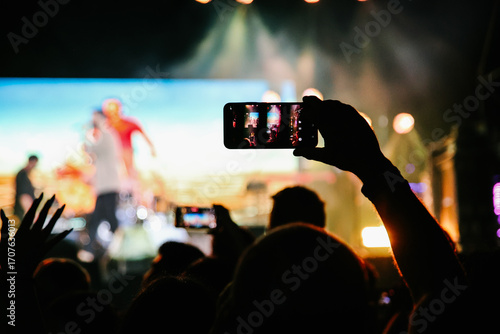 hand holding a smartphone to take a photo or record a live performance or concert with a blurred background of stage lights and bokeh