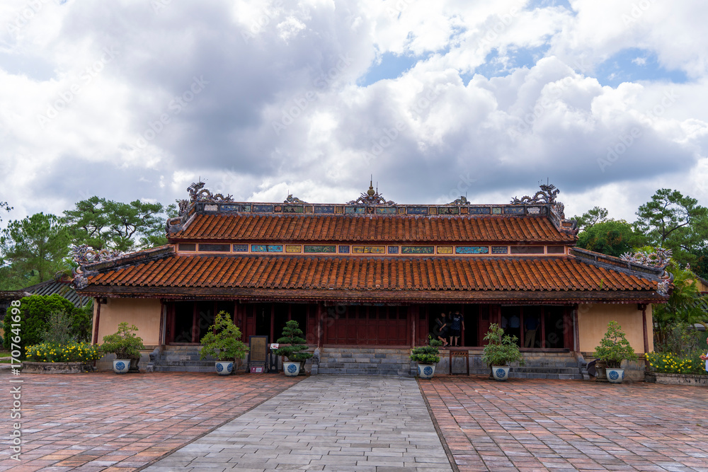 Fototapeta premium The Minh Lau pavilion, a long, single-story building with a red-tiled roof and dragon ornaments, stands centered on a brick courtyard under a cloudy sky at Minh Mang tomb, Hue.