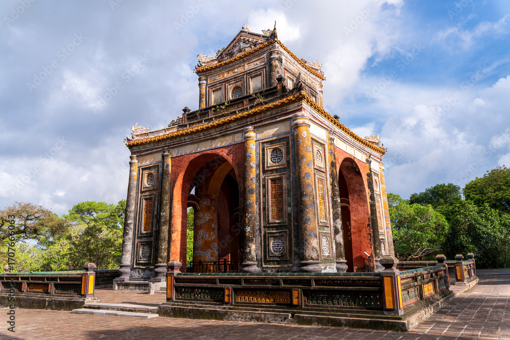 Obraz premium A weathered, ornate stone pavilion with an arched entrance houses a large stele, flanked by stone pillars and framed by trees under a cloudy sky.