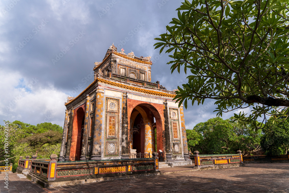 Obraz premium The Triple Gate (Tam Quan), a two-tiered, ornate structure with arches and elaborate ceramic detailing, is framed by leaves at Tu Duc tomb, Hue.