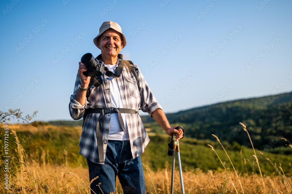 Fototapeta premium Portrait of happy and active senior hiker using camera for photographing while hiking with backpack and hiking poles in nature.