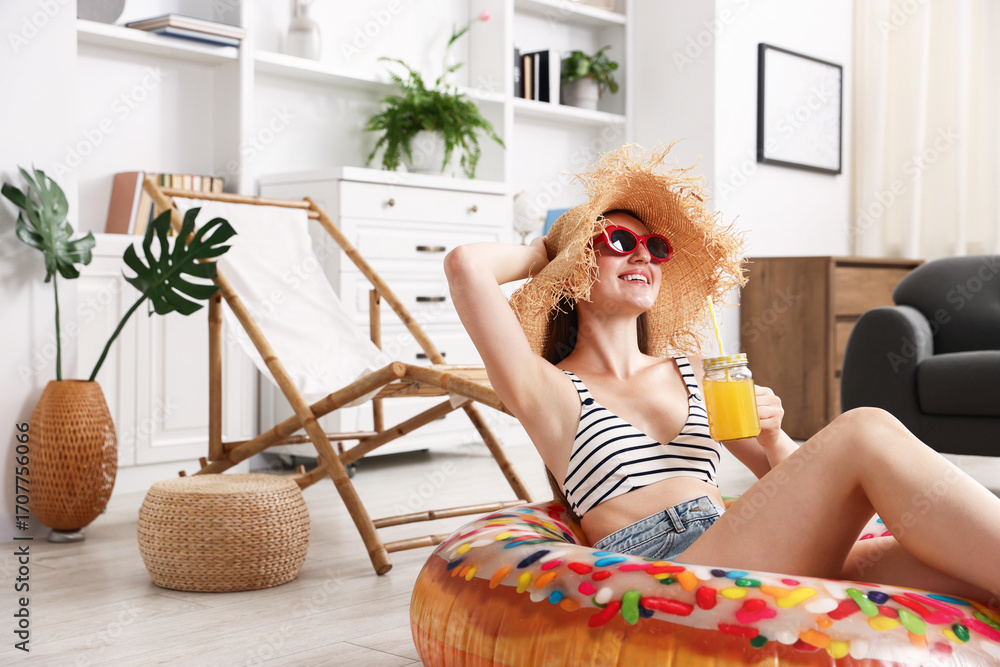 Obraz premium Happy woman in sunglasses and straw hat with mason jar of juice and inflatable ring having fun on floor at home. Dreaming about vacation at sea